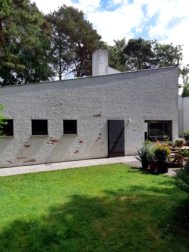 Strak gemaaid gazon bij een witte woning met dennen op de achtergrond